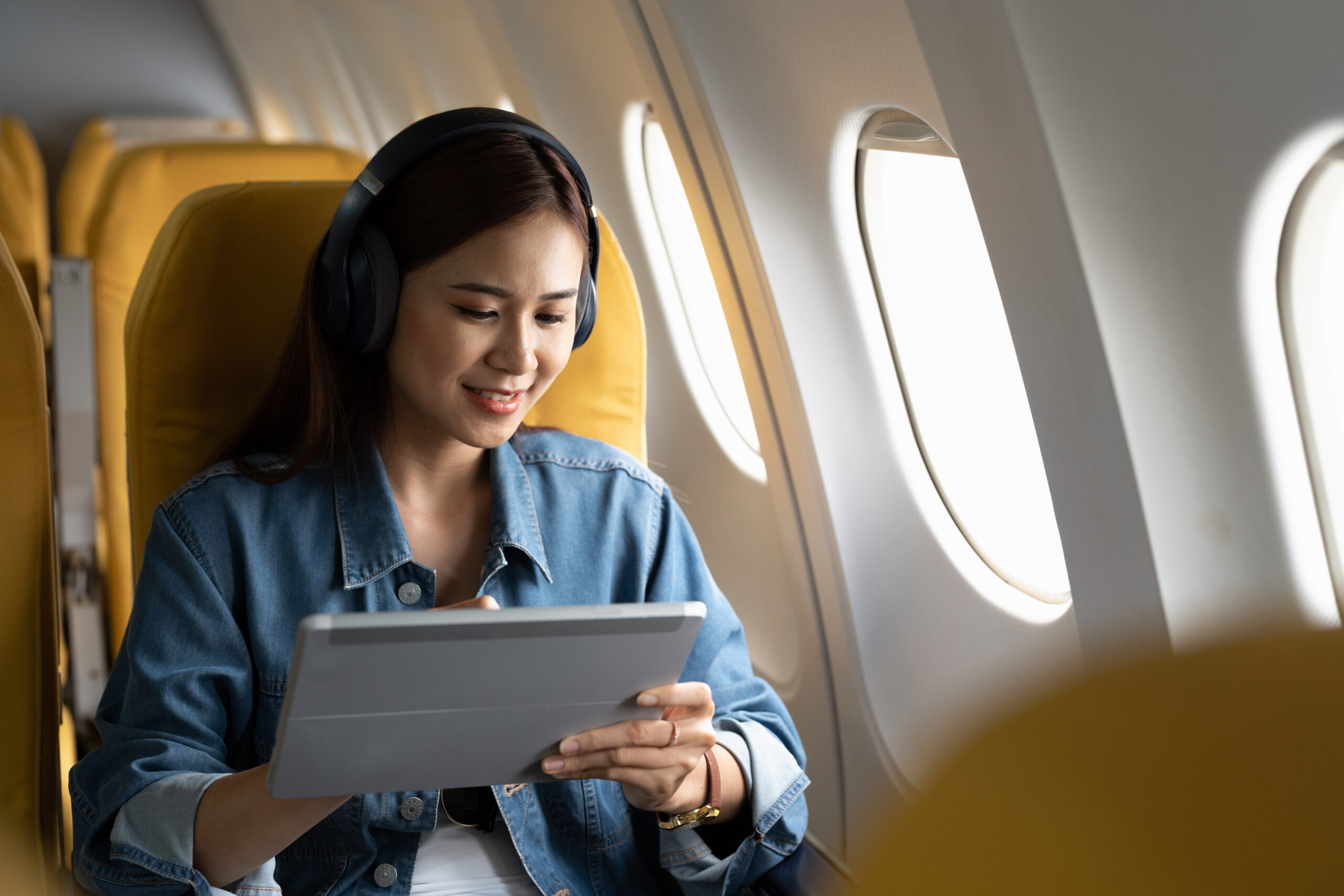 A woman wearing headphones sits in an airplane seat by the window, smiling while using a tablet. She is dressed in a denim jacket and the plane seats have yellow headrests.